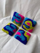This is an image of four punch needle coasters featuring an abstract freeform design of blobs and curves in hot pink, acid green, deep indigo, and teal blue. Bright Set of Coasters / Mini Rugs by dancing arms in Los Angeles, California.