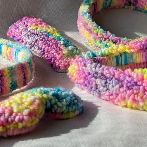 This is a close-up photo of an assortment of punch needle hair accessories. They are all made with the same bright neon rainbow yarn that ranges from yellow to teal to pink to purple.dancing arms punch needle